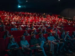 Plateia numerosa assistindo a sessão em cinema com poltronas vermelhas, iluminada pela luz da tela em ambiente escuro.