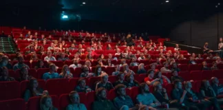 Cassinos no cinema: como Hollywood transformou as mesas de jogo em cenários inesquecíveis Plateia numerosa assistindo a sessão em cinema com poltronas vermelhas, iluminada pela luz da tela em ambiente escuro.